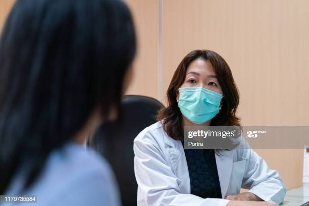 Mature doctor listening to female patient. Healthcare worker is looking at woman. Medical professional is wearing surgical mask in clinic.
